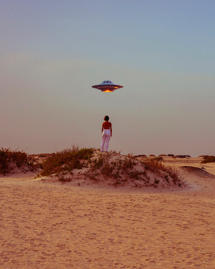Amidst the vast desert, a person captures a surreal self-portrait beneath the hovering UFO from Hey Luke, our Uber is Here! Poster. The spaceship casts a soft glow over sandy hills and serene dunes dotted with sparse vegetation.