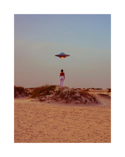 Standing on a sandy desert hill, a person gazes at a hovering spaceship in the poster Hey Luke, our Uber is Here! It features sparse vegetation and a clear gradient sky shifting from orange to blue, creating a surreal scene of wonder.