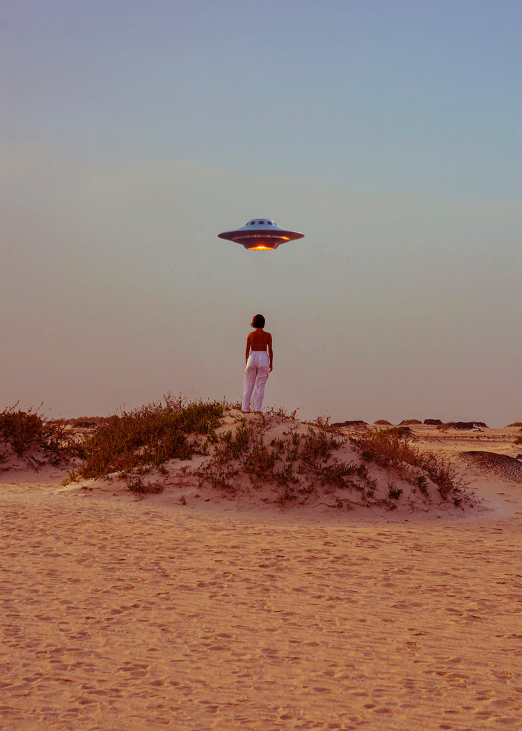 Amid the deserts expanse, a person in red and white stands on a sandy hill, capturing a self-portrait with a flying saucer above. The blue sky shifts to warm hues at the horizon, evoking collage vibes in the Hey Luke, our Uber is Here! poster.