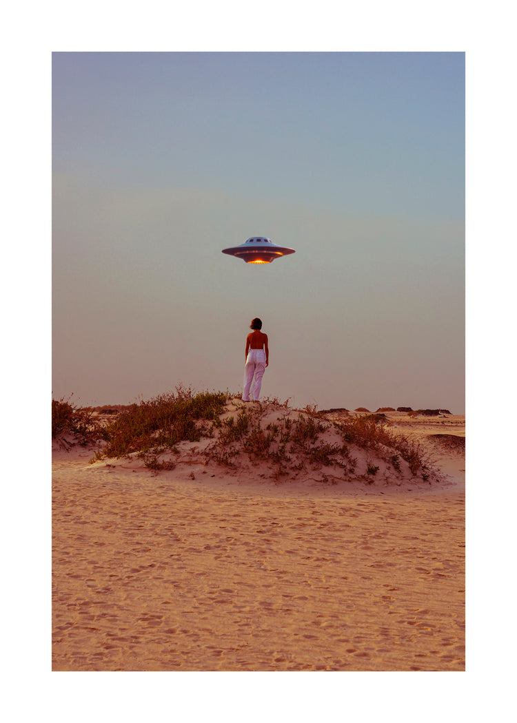 A person in white pants and a red top stands on a sandy beach, facing a hovering spaceship. Under a clear sky with blue to orange hues and bushes in the foreground, this surreal scene creates an atmosphere reminiscent of Hey Luke, our Uber is Here! poster.