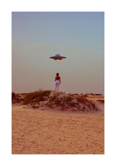 On a sandy desert dune, someone photographs themselves under the surreal scene of Hey Luke, our Uber is Here! poster. The UFO emits a soft glow against the clear sunset sky and surrounding shrubs.