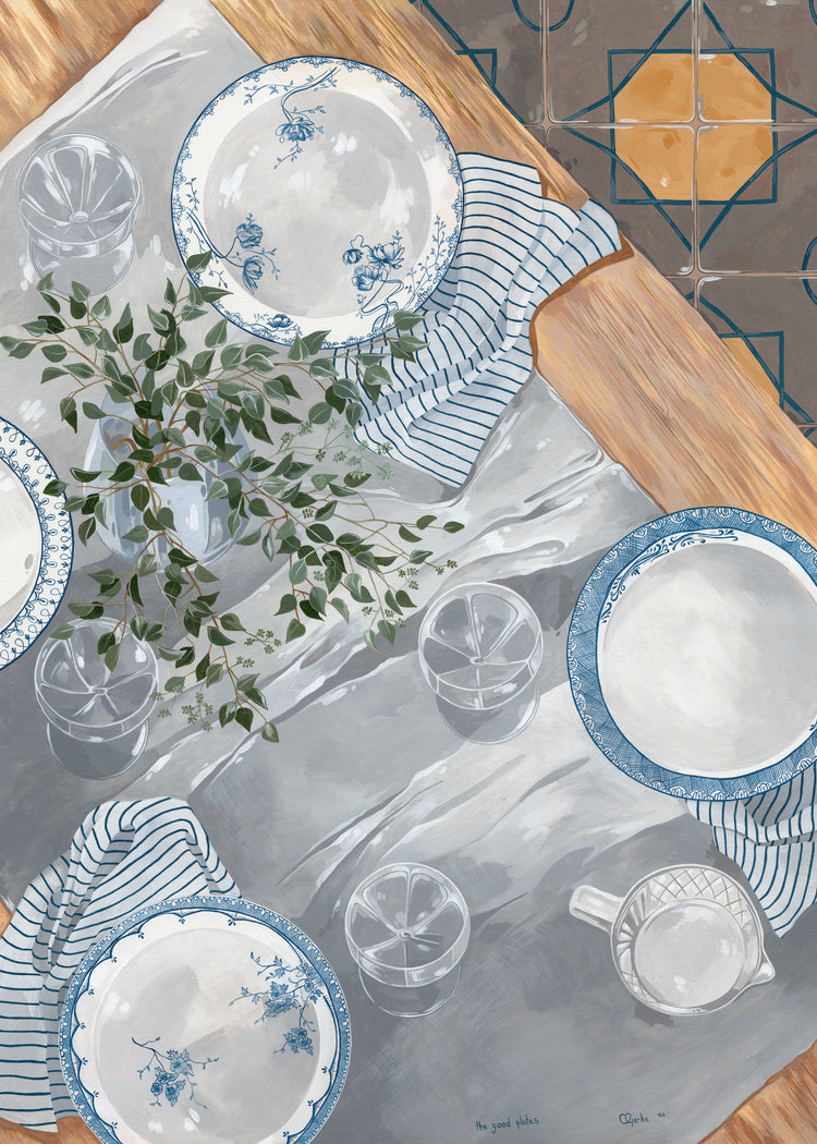 A tabletop scene featuring The Good Plates Poster with blue and white patterns, striped napkins, clear glasses, a jug on wood. A leafy green plant sits on a gray tablecloth above tiled flooring designed by Cat Gerke.