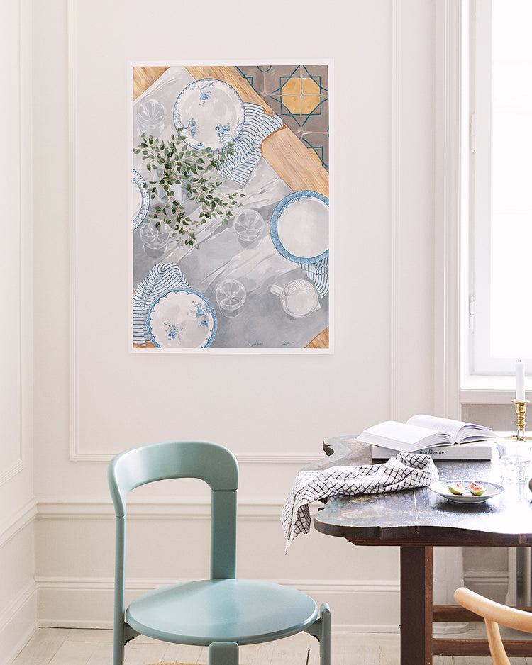 A dining room showcases a turquoise chair and dark wooden table with an open book and cloth, next to flickering candles. A white-framed artwork titled The Good Plates Poster by Cat Gerke hangs on the wall, elegantly depicting a table setting.