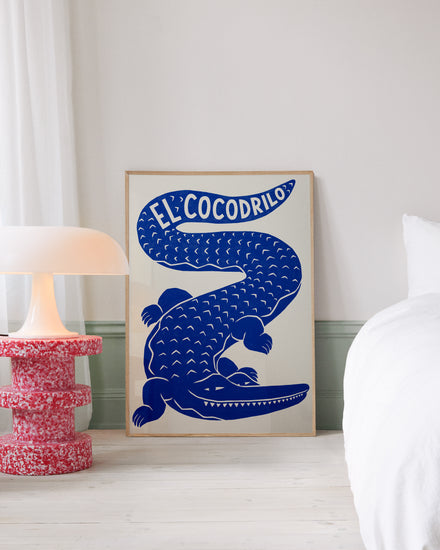 Framed poster with an illustration of a blue crocodile with the text El Cocodrilo is propped against a wall in a bedroom. The interior includes a modern white lamp on a red coffee table and a bed with white bed sheets.