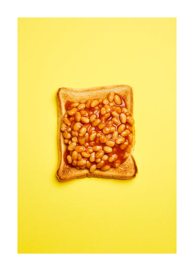 A still life photograph featuring baked beans on toast set against a vibrant yellow background. White border.