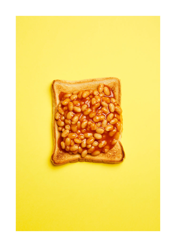 A still life photograph featuring baked beans on toast set against a vibrant yellow background. White border.