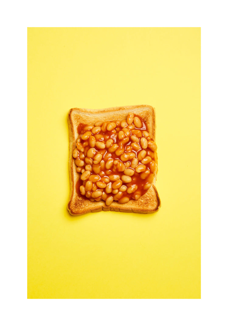 A still life photograph featuring baked beans on toast set against a vibrant yellow background. White border.