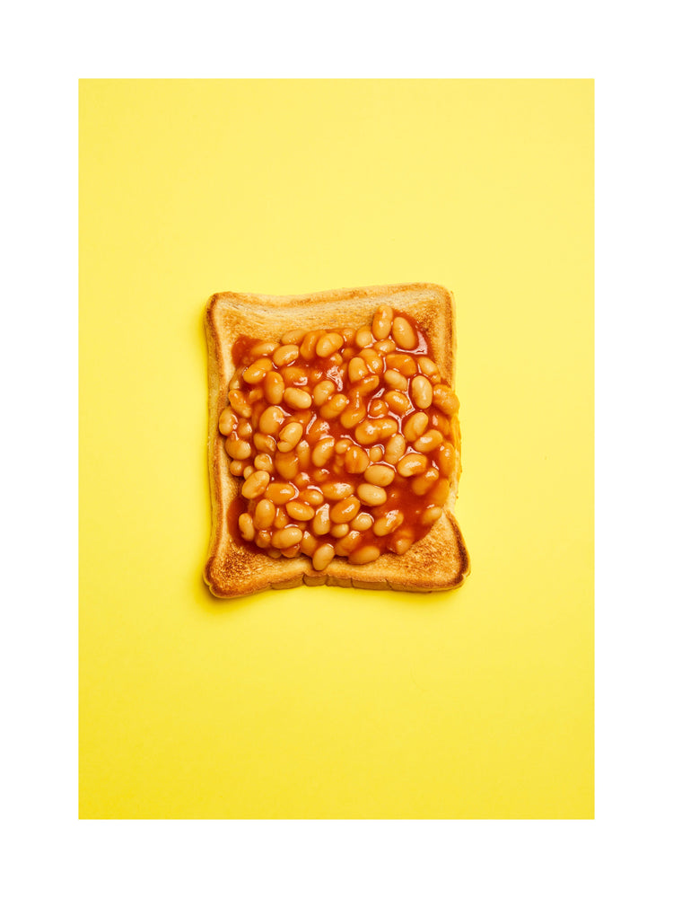 A still life photograph featuring baked beans on toast set against a vibrant yellow background. White border.