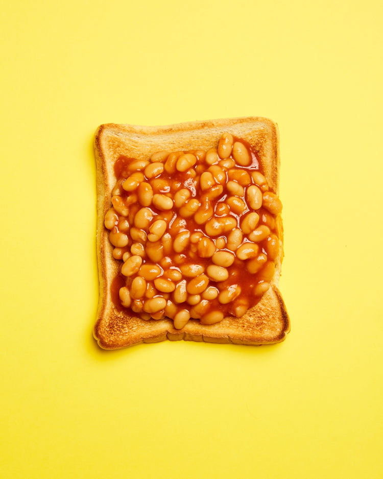 A still life photograph featuring baked beans on toast set against a vibrant yellow background.