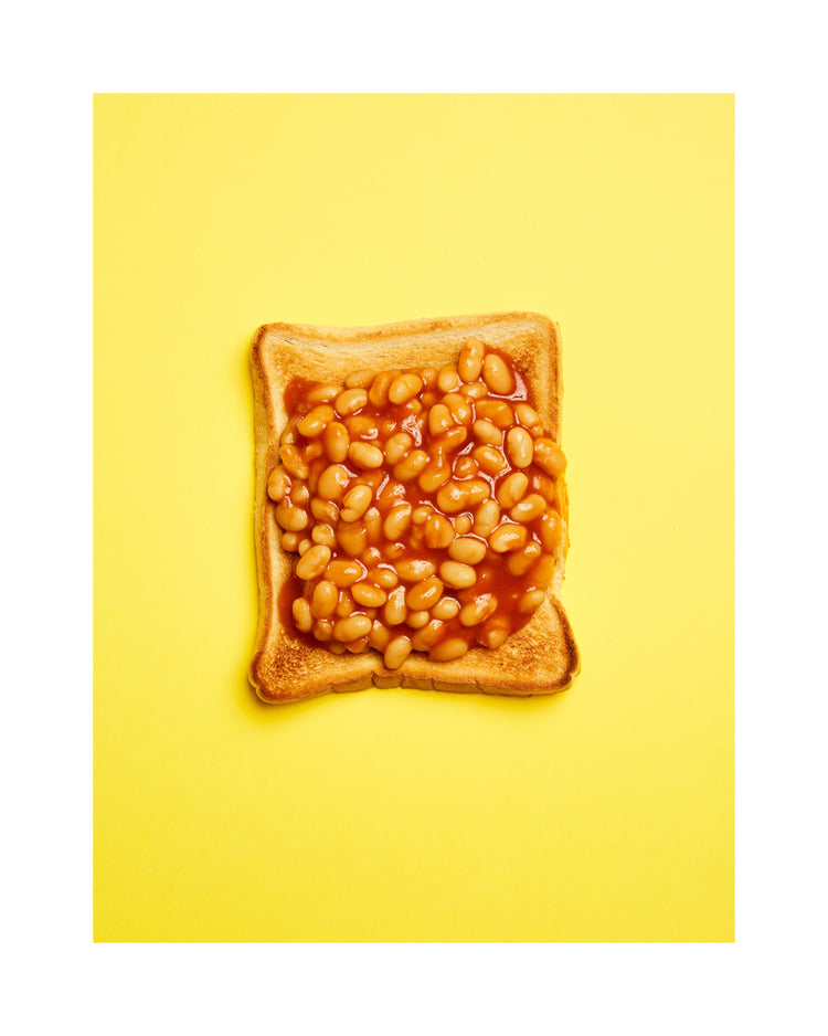 A still life photograph featuring baked beans on toast set against a vibrant yellow background. White border.