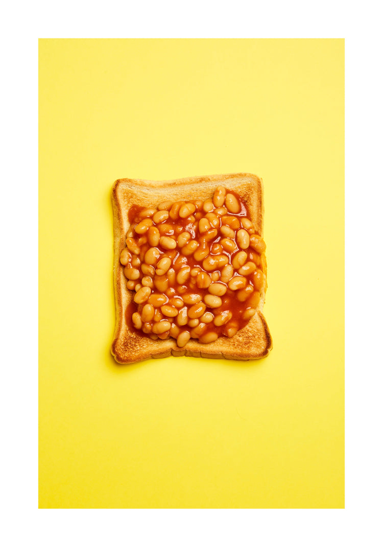 A still life photograph featuring baked beans on toast set against a vibrant yellow background. White border.