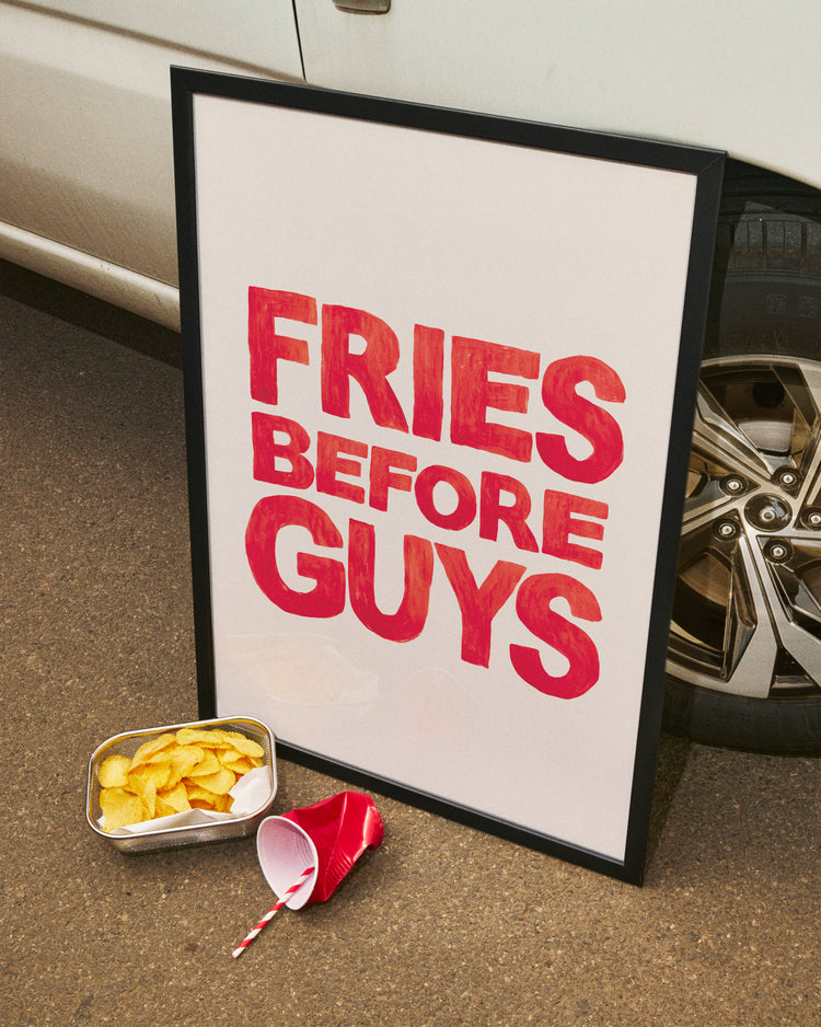 The Fries Before Guys poster, hand-painted in bold red letters, leans against a car wheel, while a foil tray full of fries and a spilled cup with a striped straw lie on the ground.