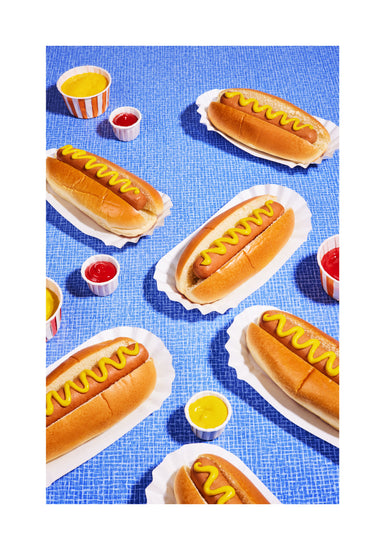 Still life photograph of perfectly grilled hot dogs adorned with mustard and served on a vibrant blue table cloth. White border.