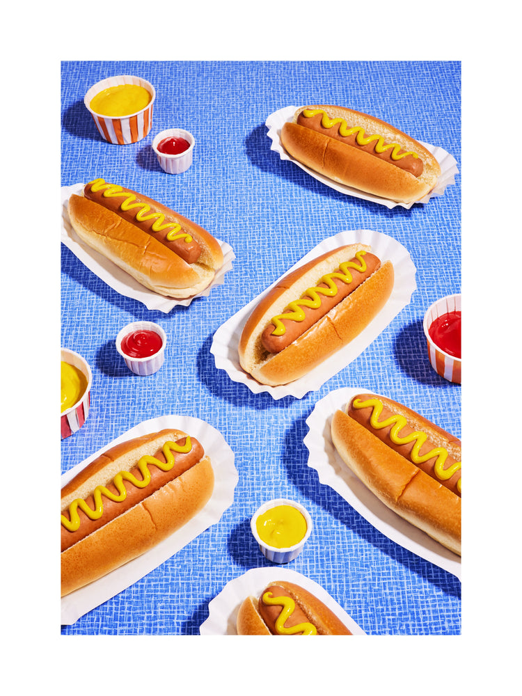 Still life photograph of perfectly grilled hot dogs adorned with mustard and served on a vibrant blue table cloth. White border.
