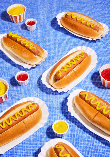 Still life photograph of perfectly grilled hot dogs adorned with mustard and served on a vibrant blue table cloth.