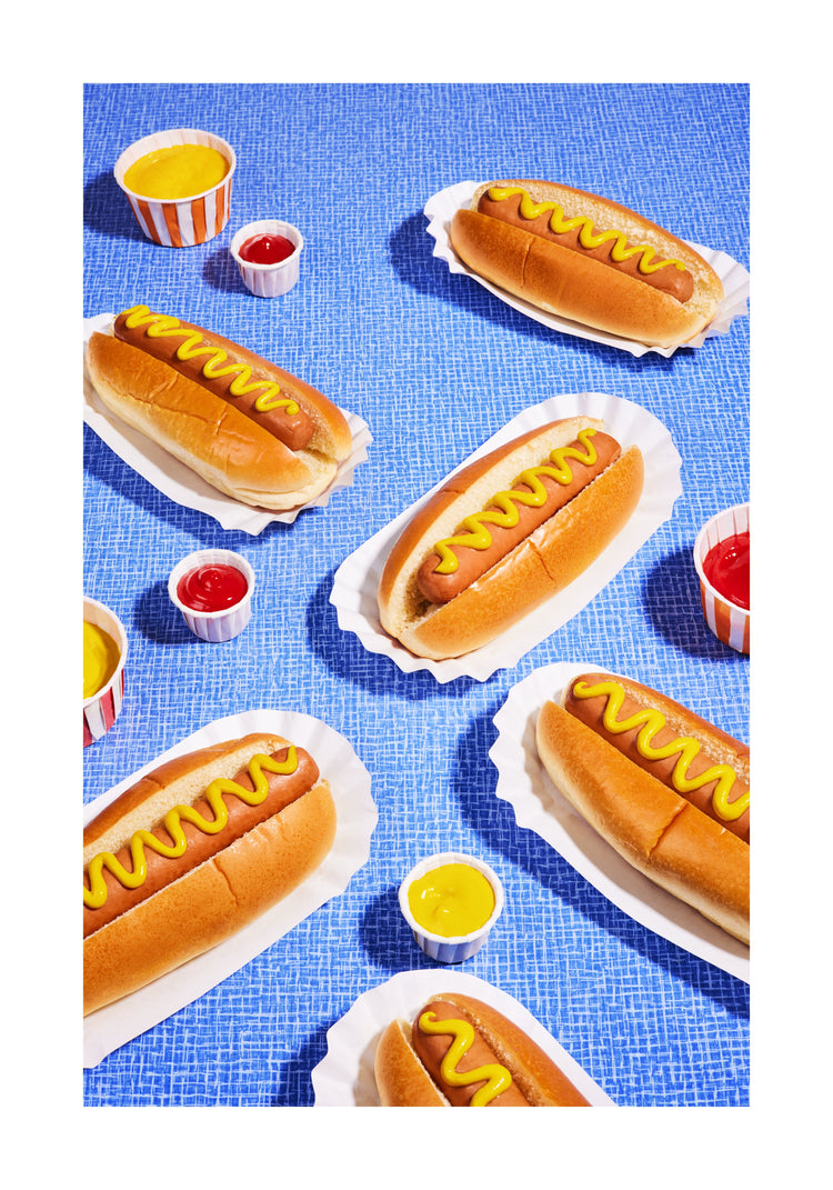 Still life photograph of perfectly grilled hot dogs adorned with mustard and served on a vibrant blue table cloth. White border.