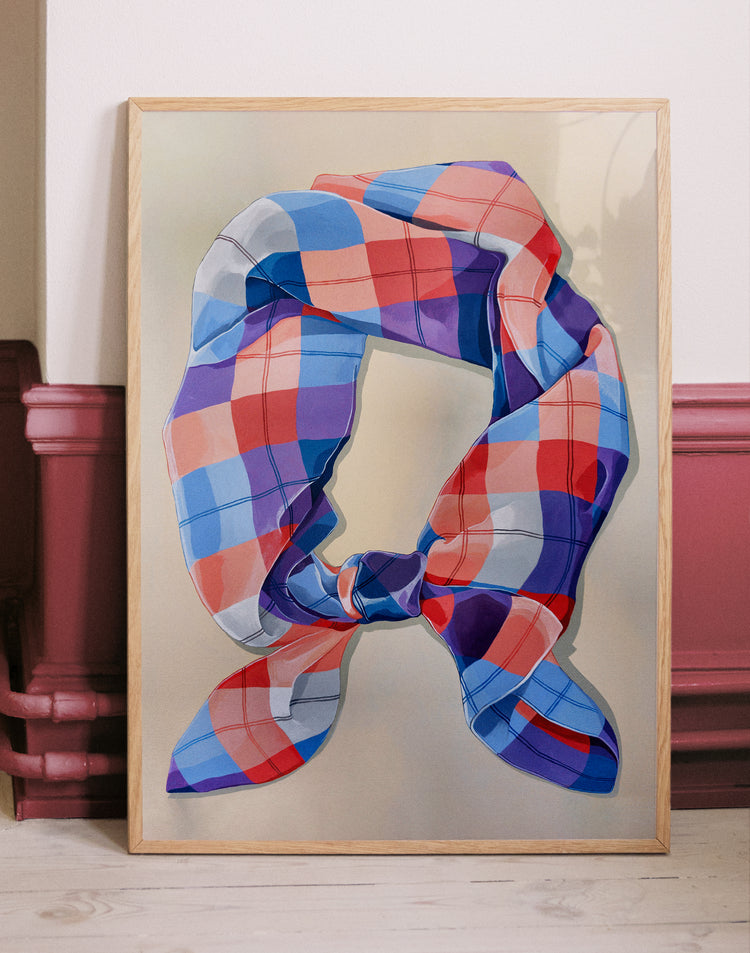 Framed A Harlequin Knot Poster by Cat Gerke, depicting a scarf in blue, red, and pink hues that appears to float magically. Exhibited on a light wooden floor against a cream and reddish-pink wall.