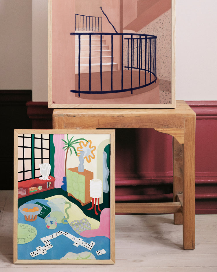The Living Room Domino Poster features two abstract paintings on a wooden chair: the top shows a staircase with a black railing, and the bottom offers an exotic room interior with plants, a rug, and dominoes against vibrant pink and maroon walls.