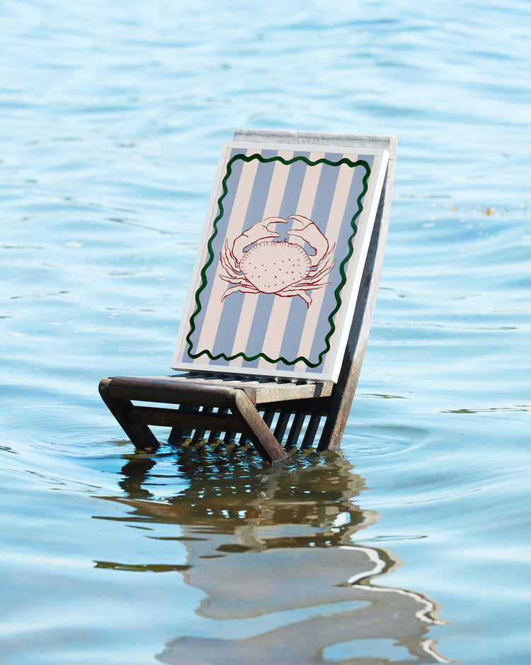A wooden beach chair, embodying coastal vibes, sits partly in serene water and showcases the Le Crabe Poster with a crab design on a striped backdrop.
