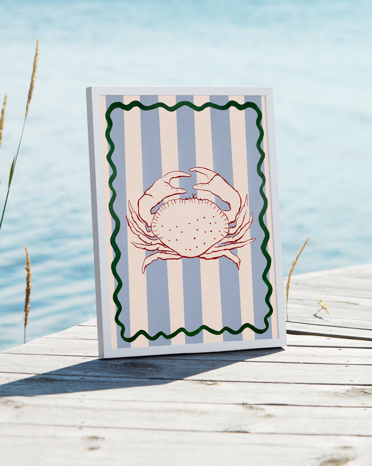 Le Crabe Poster embodies a coastal vibe with its illustration of a red crab on vertical blue and gray stripes, bordered by a green wave. Displayed against a wooden deck by water, it features tall grass gently swaying in the background.