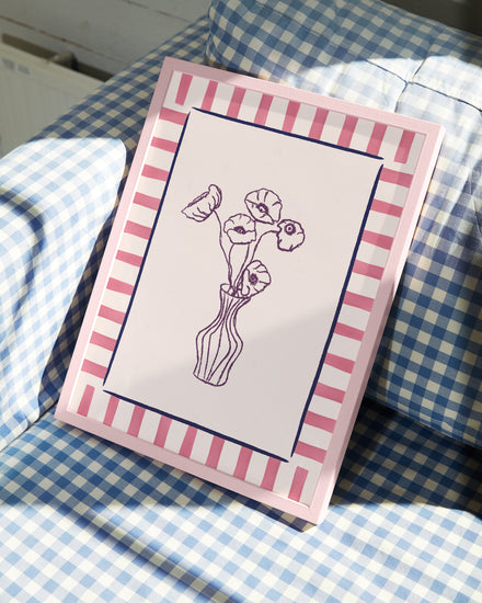 The Poppies en Vase Poster, a framed sketch of flowers in a vase with a pink and white striped border, sits on blue and white checkered fabric. Soft lighting highlights the artwork’s modern style.