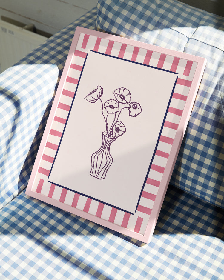 The Poppies en Vase Poster, a framed sketch of flowers in a vase with a pink and white striped border, sits on blue and white checkered fabric. Soft lighting highlights the artwork’s modern style.