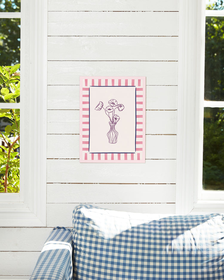 The Poppies en Vase poster with a floral motif and a pink-and-white striped border adorns a white shiplap wall. Sunlight filters through the windows, enhancing the scene, while a blue-and-white checkered couch sits comfortably below.