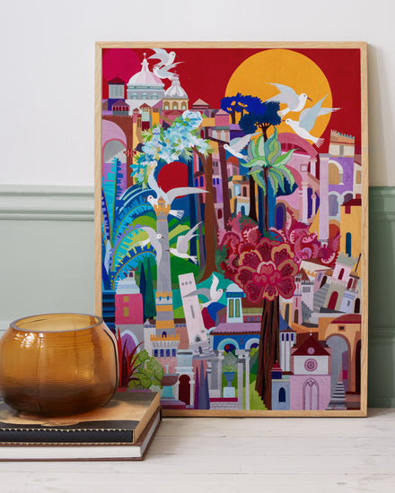The Cityscape Poster vividly captures vibrant buildings, trees, and birds against a large sun backdrop. A glass vase on stacked books rests to the left, adding charm against a dual-toned green and white wall.