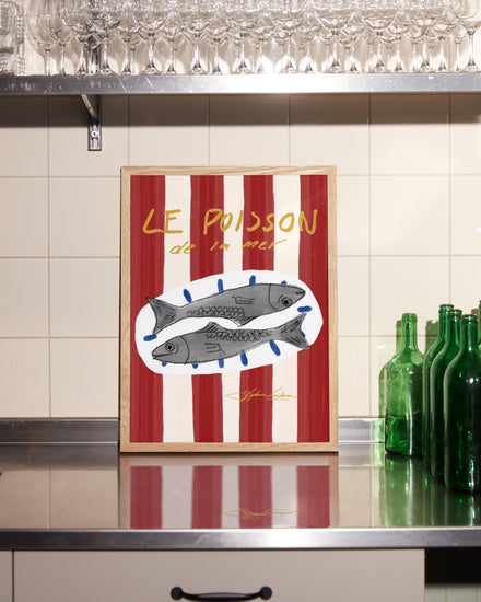 The Le Poisson Poster is a framed digital illustration capturing a coastal vibe with two fish set against a red and white striped background, displayed on a metal kitchen counter decorated with green bottles and glassware above.
