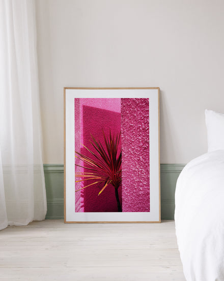 The Pink Wall in Santa Monica Poster features a vibrant pink abstract design recalling Santa Monica, with palm-like plants. It rests against a light wall next to a bed with white sheets and a transparent curtain, embodying the spirit of Californian landscape art.
