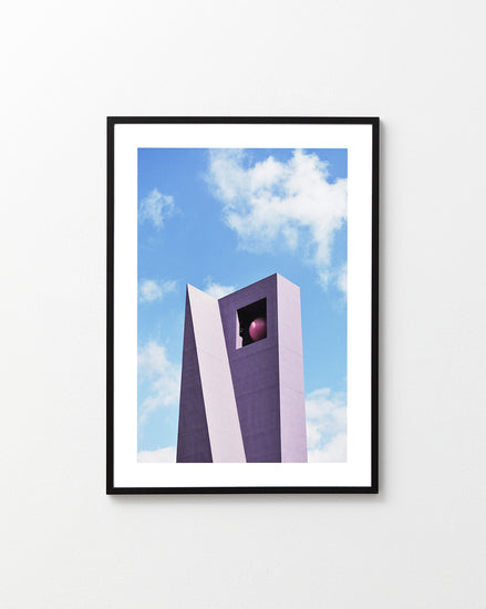 The Pershing Square Tower Poster showcases a modern, angular building with a large square opening housing a red sphere, set against a blue sky with white clouds. Created by Hanna KL, its the perfect addition to your collection.
