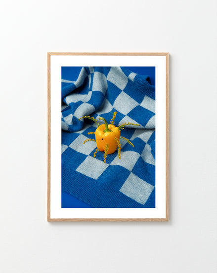 The Little Visitor Poster by Sofie Sund showcases a yellow bell pepper decorated with tiny yellow flowers and green stems, artfully arranged on a blue-and-white checkered blanket. The composition exudes a bright, fun, and vibrant poster-like vibe against the vivid blue surface.