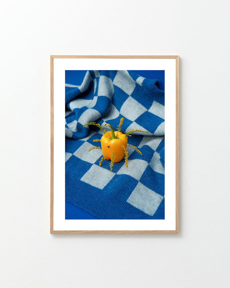The Little Visitor Poster by Sofie Sund showcases a yellow bell pepper decorated with tiny yellow flowers and green stems, artfully arranged on a blue-and-white checkered blanket. The composition exudes a bright, fun, and vibrant poster-like vibe against the vivid blue surface.