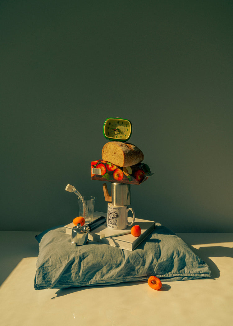 A still life featuring various breakfast elements including a slice of bread, apple juice, coffee mug, teapot, with a pillow underneath and an alarm clock on top.