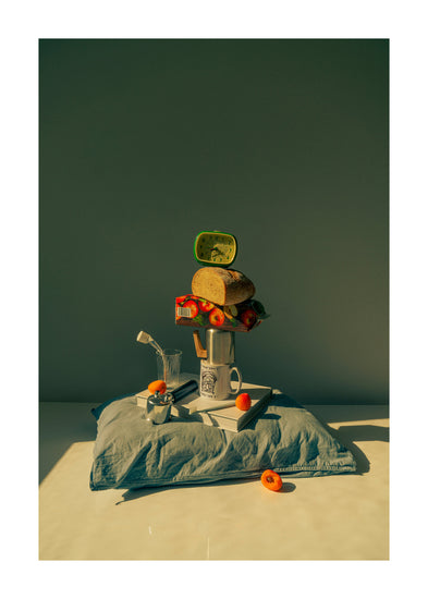 A still life featuring various breakfast elements including a slice of bread, apple juice, coffee mug, teapot, with a pillow underneath and an alarm clock on top.