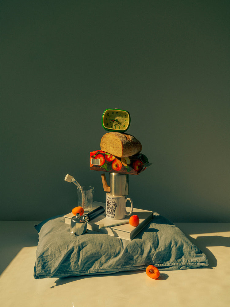 A still life featuring various breakfast elements including a slice of bread, apple juice, coffee mug, teapot, with a pillow underneath and an alarm clock on top.