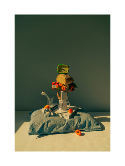 A still life featuring various breakfast elements including a slice of bread, apple juice, coffee mug, teapot, with a pillow underneath and an alarm clock on top.