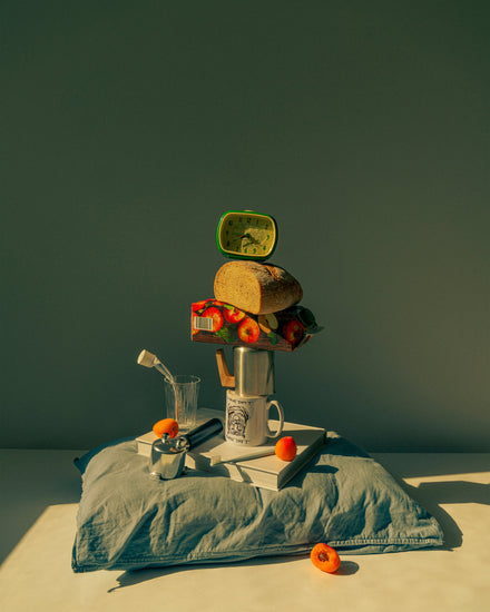 A still life featuring various breakfast elements including a slice of bread, apple juice, coffee mug, teapot, with a pillow underneath and an alarm clock on top.
