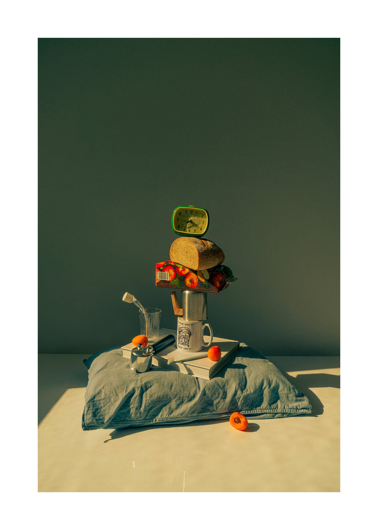 A still life featuring various breakfast elements including a slice of bread, apple juice, coffee mug, teapot, with a pillow underneath and an alarm clock on top.