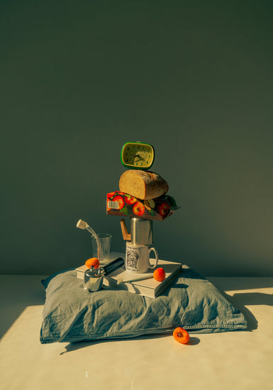 A still life featuring various breakfast elements including a slice of bread, apple juice, coffee mug, teapot, with a pillow underneath and an alarm clock on top.