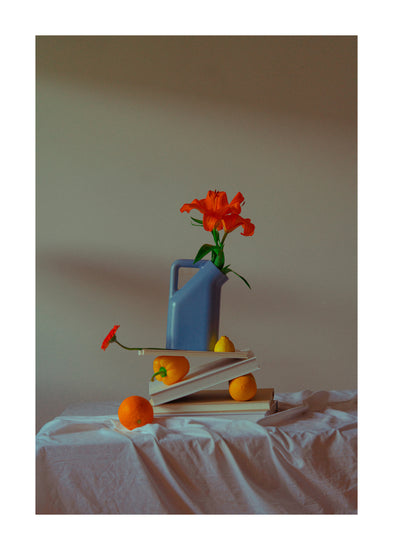 The Hanging in There Poster showcases a still life with a blue watering can, orange flower, books, and fruit on a cloth-covered table. The vibrant colors contrast like Berlins winter scenes, with soft lighting casting gentle shadows.