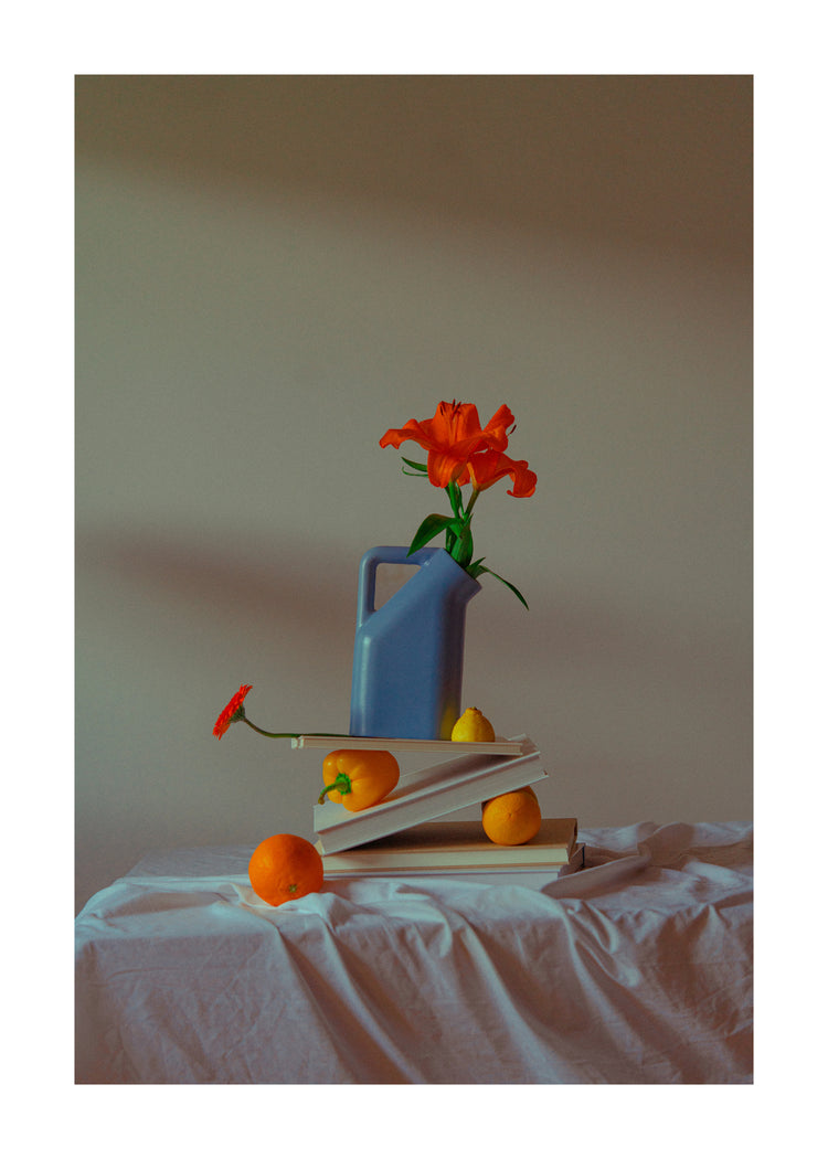The Hanging in There Poster showcases a still life with a blue watering can, orange flower, books, and fruit on a cloth-covered table. The vibrant colors contrast like Berlins winter scenes, with soft lighting casting gentle shadows.