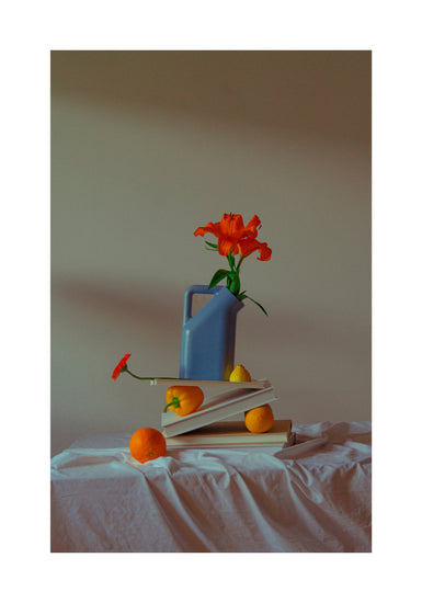 The Hanging in There Poster depicts a vibrant still life with a blue watering can and orange flowers on stacked books, set against Berlins colorful backdrop. Oranges, lemons, and a red flower are artfully arranged on a draped white cloth in this lively winter scene.