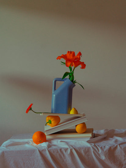 The Hanging in There Poster from Berlin presents a vibrant still life featuring a purple jug with orange lilies on books, accompanied by an orange, two lemons, and a red flower on a white cloth. The neutral backdrop emphasizes the vivid hues, capturing winters longing for brightness.