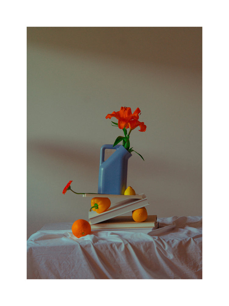 A Hanging in There Poster shows a blue vase with orange flowers on a book stack draped in white cloth. Oranges and a lemon scattered around cast soft shadows on the muted background, evoking vibrant winter colors of Berlin.