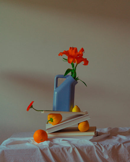The Hanging in There Poster showcases a still life with a blue pitcher holding a red flower, resting on books. An orange, yellow pear, and daisy surround them, their vibrant colors contrasting the muted background and evoking Berlins winter warmth.
