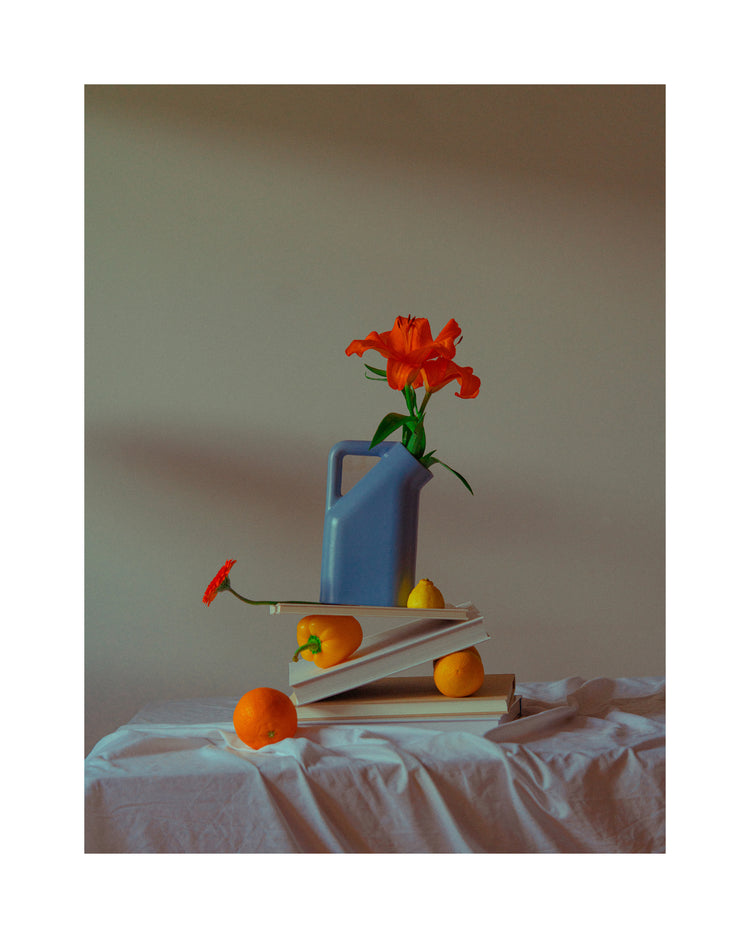 The Hanging in There Poster features a still life with a blue pitcher holding an orange flower on books, capturing Berlins vibrant colors. Oranges and a lemon rest on a white tablecloth, all set against a plain wall for a minimalist winter scene.