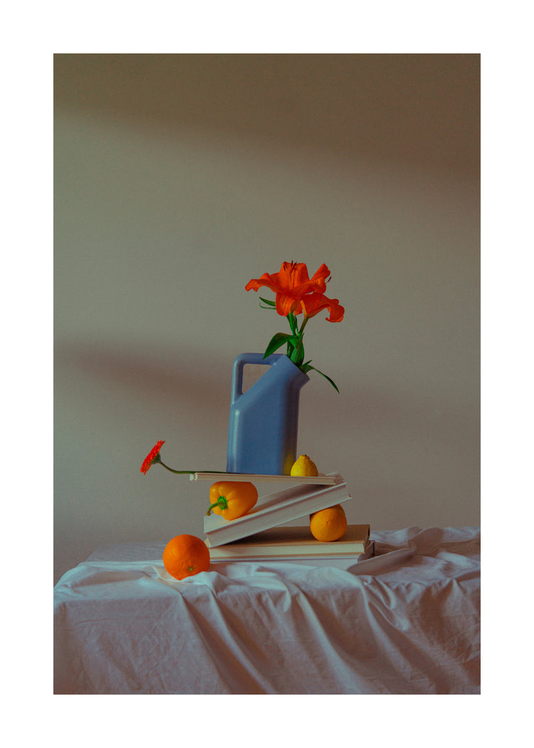 The Hanging in There Poster shows a blue pitcher with an orange flower on stacked books, alongside an orange, lemon, and a red flower leaning on the orange against a backdrop resembling the serenity of a Berlin winter.