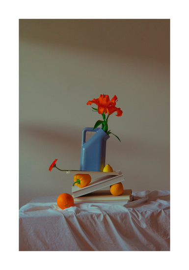 The Hanging in There Poster showcases a still life scene with a blue watering can, an orange flower, stacked books, oranges, and a pear on white cloth against a neutral Berlin backdrop, capturing vibrant winter tranquility.