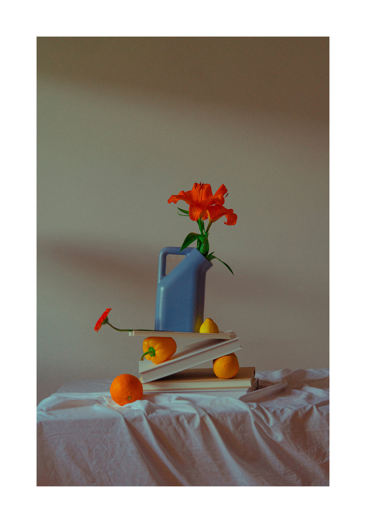 The Hanging in There Poster showcases a still life scene with a blue watering can, an orange flower, stacked books, oranges, and a pear on white cloth against a neutral Berlin backdrop, capturing vibrant winter tranquility.